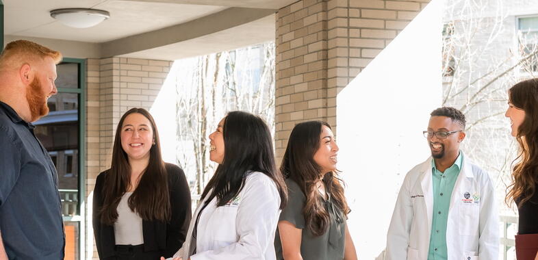 students in and out of white coats, conversing outside in the shade of a building