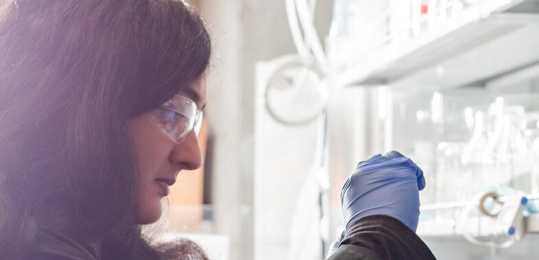 A lab worker with safety glasses works, holding up a sample to the light of a window