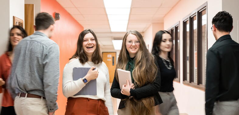 two girls look at camera and smile while holding their laptops in a hallway. people walk past them.