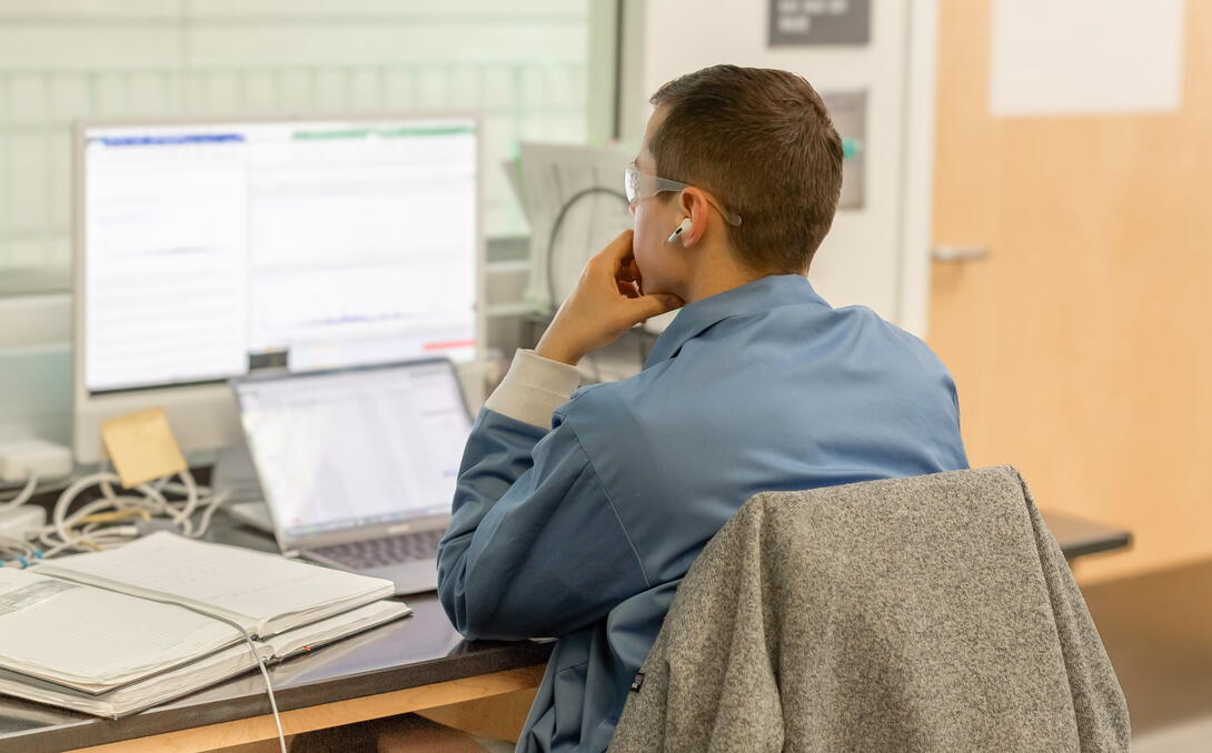 man in lab coat works on a series of computer monitors, looking thoughtful