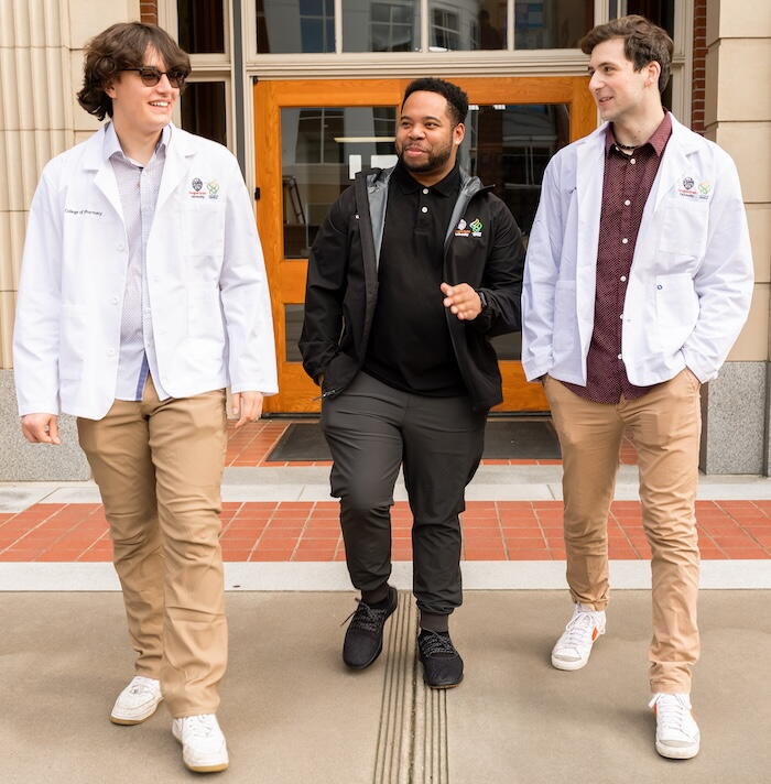 Three students smile and speak with one another outside of the front of the college of pharmacy building